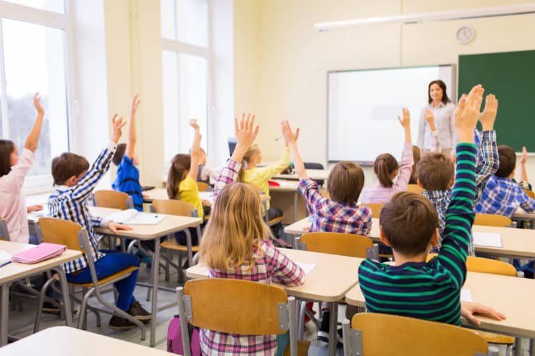 group of school kids with teacher sitting in classroom and raising hands