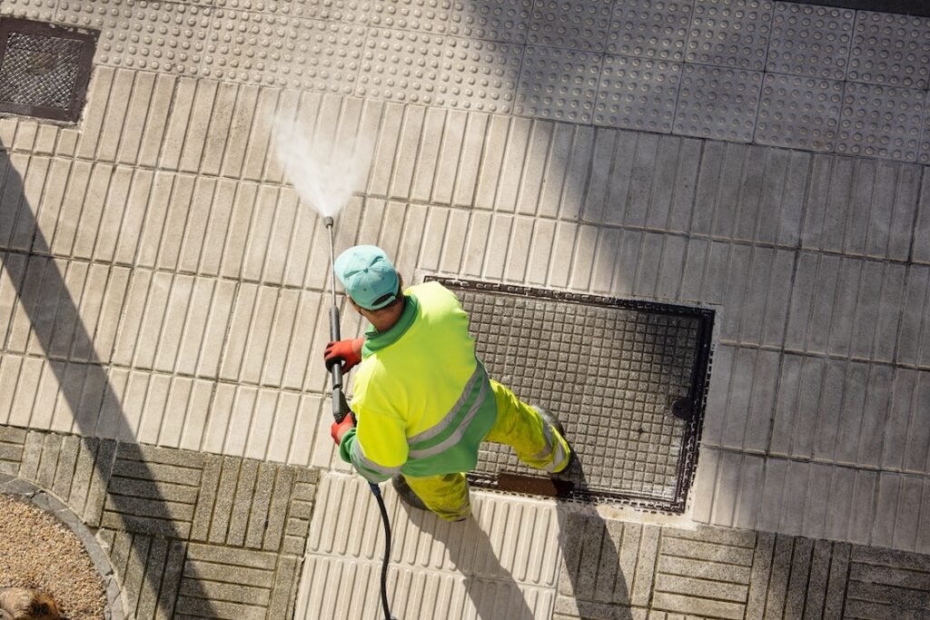 Worker cleaning a street sidewalk with high pressure water jet machine on sunny day.